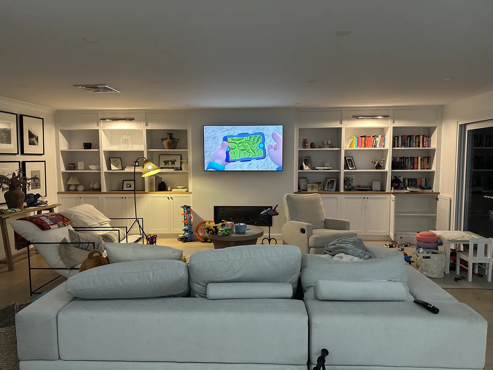 Family living room in the evening with built-in shelving, a wall TV and toys, before